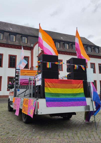 Hochformat-Foto: Ein Kleinlaster von schräg hinten steht auf dem Karlaplatz in Heidelberg. Die Ladefläche ist mit vielen Pride-Flags geschmückt, aufgesteckt sind vier Lesbian-Pride-Flags und eine Trans-Flagge. Große Lautsprecher stehen auf der Ladefläche. Man kann sich das Bild sehr laut vorstellen!