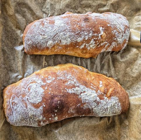 zwei Ciabattabrote, goldbraun gebacken und mit Mehl bestreut, auf hellem, etwas krumpeligem Backpapier. das obere ist deutlich kleiner als das untere