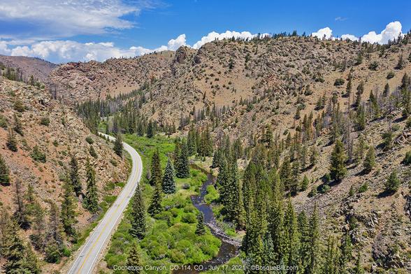 Aerial photo looking up a narrow, shallow canyon. A large stream winds down the bottom with lush green shrubs and tall conifer trees on either bank. A two lane highway follows the left side of the canyon. The canyon is formed by hills of rock and soils, dry and mostly vegetation free. Blue sky with some clouds.
©BosqueBill.com