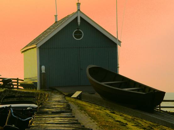 Querformat: Irgendwo in Norwegen. Ein blaues Bootshaus mit einem kleinen runden Fenster, ein dunkles Boot davor, das auf einer kleinen Rampe liegt, Steinmauer im Vordergrund, orangefarbener Himmel im Hintergrund.