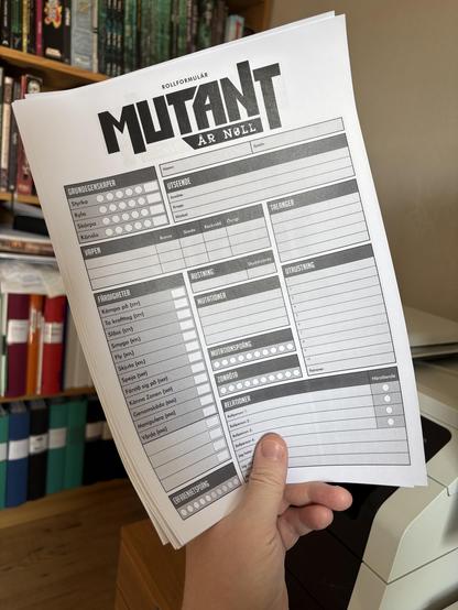 Fotografi av en bunt med rollformulär till rollspelet Mutant År Noll som hålles upp framför en datorprinter. I bakgrunden syns bokhyllor med rader av pärmar och rollspel.