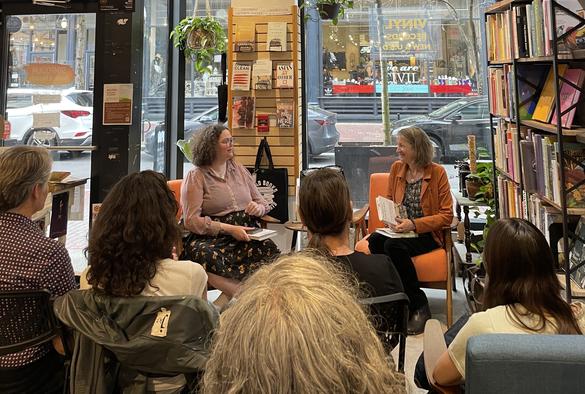 Photograph of author Kirsten Menger-Anderson in conversation about her novel THE EXPERT OF SUBTLE REVISIONS with writer and visual artist Tanya Young at Symposium books in Providence, RI. In the foreground is the audience, with bookshelves to the right and a floor to ceiling window in the background.