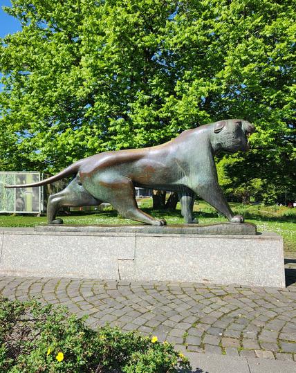 Eine große Bronze-Vollplastik von der Seite aufgenommen.
Das Tier steht auf einem Steinsockel. Der Körper ist angespannt, die 4 Läufe deuten eine nach vorne gehende Bewegung an. Das Maul ist offen, der Kopf erhoben.