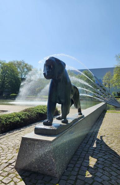 Die gleiche Tierplastik von unten-vorne. Jetzt sieht man das aggressiv-offene Maul mit 2 Reißzähnen. Im weiteren Hintergrund eine hohe Fontäne mit mehreren einzelnen und langen Strahlen, die seitlich aus einer Leitungsöffnung in ein flaches Wasserbecken schießen. Der Himmel darüber ist strahlend blau.