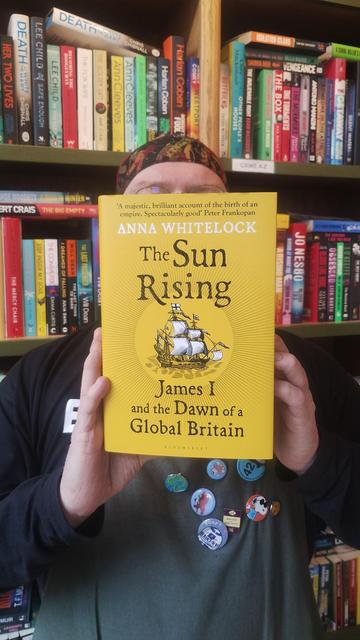 Bookseller Joe holding up a hardback copy of The Sun Rising, by Professor Anna Whitelock, a bright yellow cover with a period sailing vessel on it, in the Edinburgh Bookshop