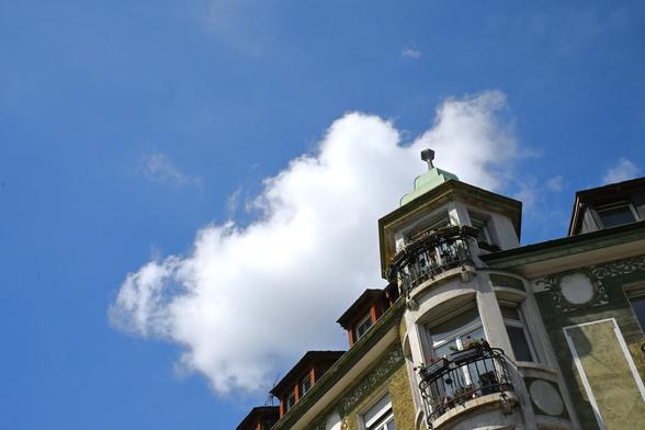 The image features a building with a distinctive architectural style. The focus is on the upper part of the structure, showcasing a rounded turret with a green dome. There are balconies adorned with plants, and the building has a weathered appearance with decorative elements on its facade. The sky is bright blue with a few fluffy white clouds, providing a contrasting backdrop to the building.
