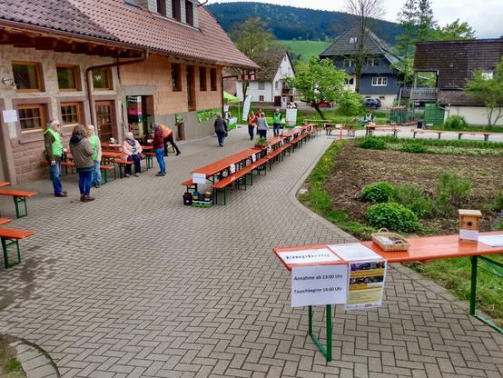 Zu Beginn stellen wir die roten Tische und Bier-Bänke auf, die hier auf einem gepflasterten Weg zus ehen sind. Links ist das Haus des Biohofes, rechts ein unbepglanztes Beet, im Hintergrund Häuser des Dorfes und ein bewaldeter Hügel, der Teil des Schwarzwaldes ist.
