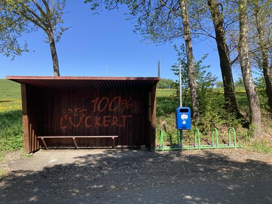 Die Bushaltestelle in Lückert. Sie ist aus Holz. Ich habe sie frontal fotografiert. Daneben links hängt ein blauer Mülleimer und dahinter steht ein knallgrüner Fahrradständer. Der Himmel st von kräftigem Blau. Einige Bäume rechts und links.