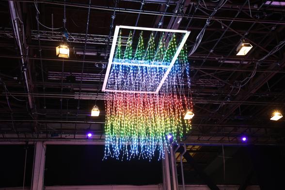 A 3D LED matrix out of LED stripes hanging down equally spaced from a wooden rectangle. It is showing rainbow colors.