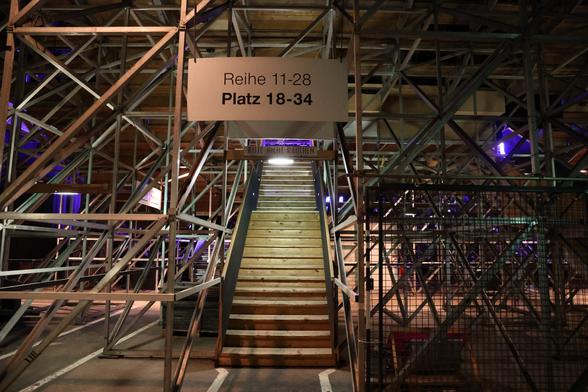 Lots of scaffolding below a tribune. A wooden stair leads into the seating area, framed by big signs "Row 11-28, Seat 18-34" and "No smoking please"