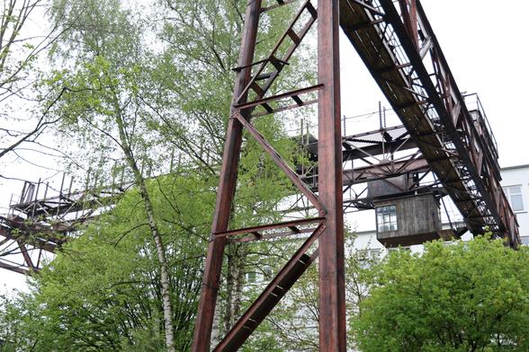 Some rusty industrial scaffolding outside, going up, forwards and left. Probably for transportation or loading of goods? Green trees grow in between.