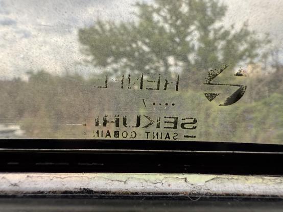 Foto de la parte inferior de una ventana con la parte que se abre de forma que se tumba. Se ve el logotipo antiguo de RENFE con la galleta y el texto “RENFE” y abajo pone el logo del fabricante “Sekurit Saint-Gobain”.

De fondo se ve unos árboles.