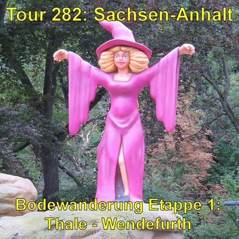 Eine große Plastikfigur einer Hexe in leuchtend pinkem Kleid und spitzem Hut steht auf einem künstlichen Felsen. Die Figur hat blonde Haare, ausdrucksstarke blaue Augen und hält beide Arme weit ausgestreckt. Das Kleid hat weite Ärmel und einen Schlitz auf der Vorderseite. Im Hintergrund sind hohe Bäume mit dichtem grünen Laub zu sehen.