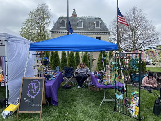 Photo of my booth at Fairyville in Noblesville, Indiana. I’m at the center underneath a blue tent. On the sides are two tables with purple tablecloths. A sign on the left reads “Melly’s Distractions” in white. Above it is a purple logo with a swooping M and D.