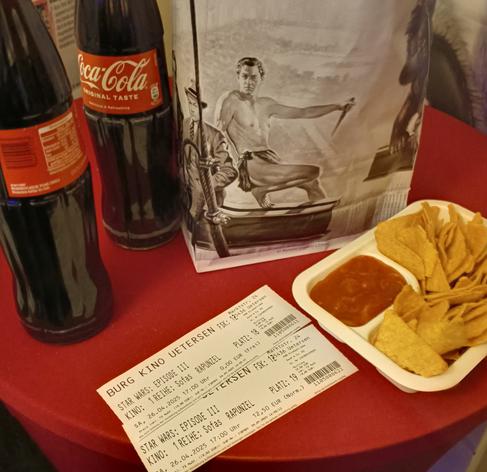 Auf einem runden Tisch mit roter Decke stehen zwei Flaschen Cola, eine Tüte Popcorn, eine Portion Nachos und zwei Eintrittskarten