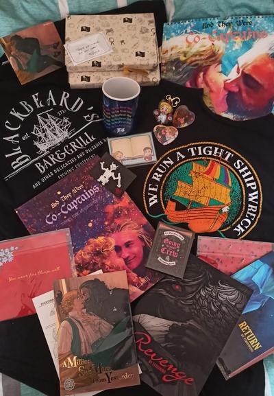 A miscellany of items made by fans of Our Flag Means Death. There are two t-shirts, a calendar, an artbook, a mug, a few doujinshi, a deck of inspirational cards, a notepad, a glasses cleaning cloth based on the piece of red fabric, and two rectangular objects wrapped in OFMD-themed wrapping paper.