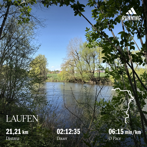 Blick über einen Fluss (Saale), eingerahmt von Frühlingssträuchern und sonnenbeschienenen Bäumen, blauer Himmel und am Horizont ist eine Siedlung (Schellsitz) mit einer Kirche erkennbar
