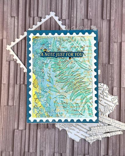 A greeting card in blue and green tones with gold foil accents. The main feature of the card looks like an oversized postage stamp. The background is blue with layers of different textures in green and gold. The greeting reads "A note just for you."