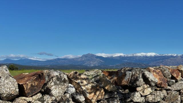 A view towards the snow tipped mountains of the Sierra de Madrid. In the foreground a stone wall runs across the picture made up of stones coloured grey and brown.