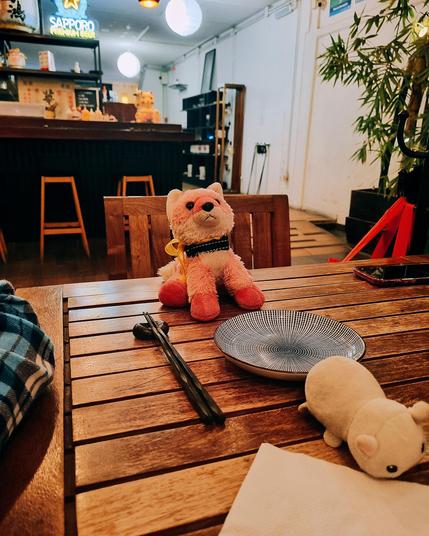 The image shows a wooden table in what appears to be a Japanese restaurant. There are two stuffed animals, one pink and one white, arranged on the table as if they are dining. A plate and chopsticks are placed in front of the pink stuffed animal. The restaurant is empty and dimly lit.