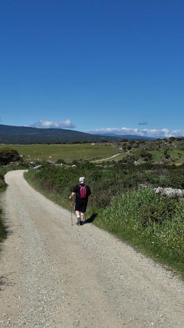 My partner walks down a sandy path which winds through the green hill landscape. There are banks of earth on either side as well as hedges. The sky is nearly cloudless and bright vibrant blue.