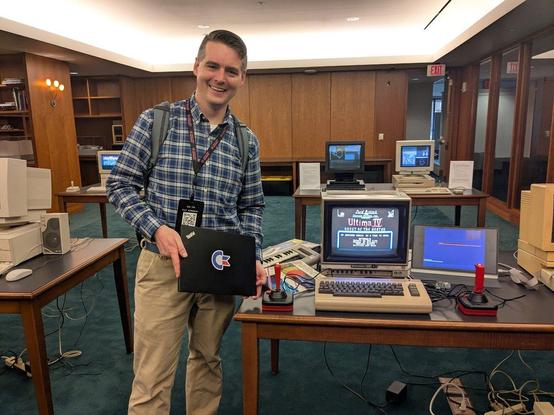 A person (me) standing in front of a Commodore 64 display holding a laptop with a Commodore logo sticker. A Next workstation and Amiga 3000UX is in the background.