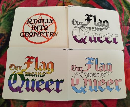 4 stickers with different designs: A "Really into Geometry" one with a red pentagram in the background. Three "Our Flag means Queer" logos with the letters coloured as different flags: ace, gay and trans.