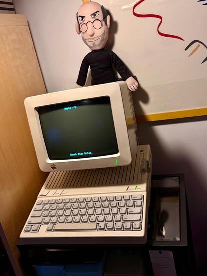 Apple //c setup on a small shelf with Steve Jobs doll on top of the CRT