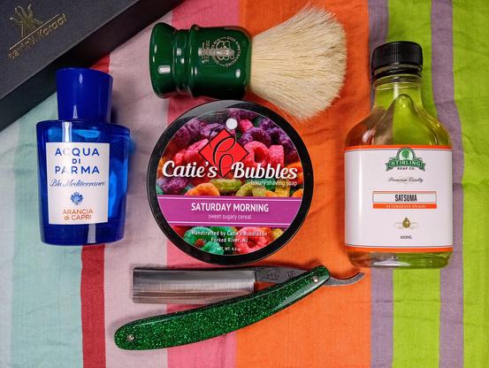 (Le Chat Mistral description)

Thank you for the correction. Here's the revised description:

This image shows a collection of grooming items arranged on a colourful striped cloth. There's a blue bottle of Acqua di Parma cologne labelled "Arancia di Capri" on the left. Next to it is a round container of Catie's Bubbles shaving soap, which has a vibrant label featuring colourful cereal pieces and is titled "Saturday Morning." To the right, there's a green bottle of Stirling Soap Co. aftershave splash labelled "Satsuma." Above these items, there's a shaving brush with a deep green handle. A green-handled straight razor lies below the items, adding a classic touch to the arrangement.o
