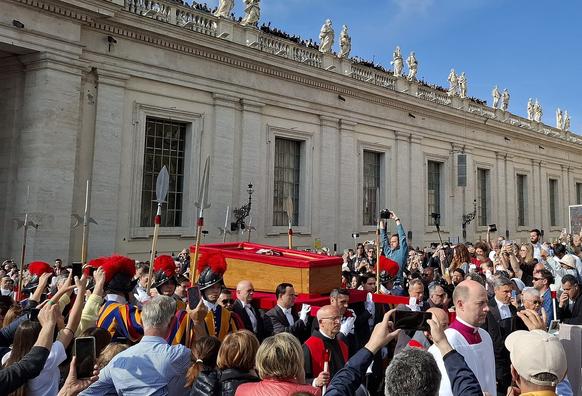 Al centro le guardie svizzere, armate di lance, scortano la bara del papa trasportata da alcuni uomini in giacca e papillon. Intorno una folla di persone, molte scattano foto. SUllo sfondo un edificio del Vaticano.