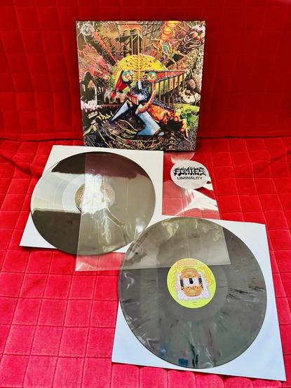 A colorful, psychedelic-themed vinyl album set displayed on a bright red quilted background. The album, titled "Liminality" by the band FOMIES, features vibrant, chaotic artwork with surreal, science-fiction elements, including a robotic figure and abstract machinery. Two black and smoky translucent vinyl records are placed in white inner sleeves, one partially removed and the other fully visible. A circular sticker with the band’s name "FOMIES" and the word "LIMINALITY" in black, bold letters is also included.