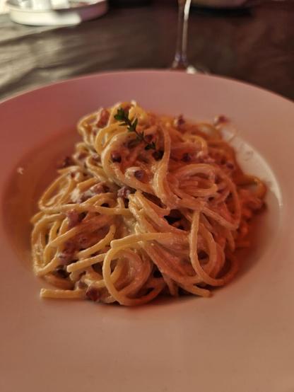 Das Bild zeigt eine Portion Carbonara auf einem weißen Teller. Die Spaghetti sind in einer cremigen Sauce mit kleinen Stückchen von Speck vermischt. Ein kleiner Zweig von Thymian ist als Garnitur auf der Pasta platziert. Der Teller steht auf einem dunklen Tisch, und im Hintergrund ist ein Glas mit Wein zu sehen. Die Beleuchtung ist warm und einladend, was die Farben der Pasta und der Sauce hervorhebt.
