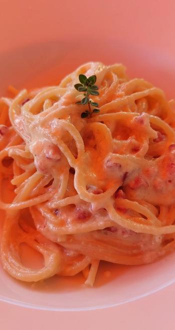 Das Bild zeigt eine Portion Spaghetti Carbonara auf einem weißen Teller. Die Spaghetti sind in einer cremigen, orangefarbenen Sauce angerichtet, die wahrscheinlich aus Eiern, Schinken und Parmesan bestehen könnte. Die Sauce bedeckt die Nudeln vollständig und hat eine glänzende, reichhaltige Textur. Einige kleine Stücke von Schinken sind in der Sauce sichtbar. Oben auf der Portion befindet sich ein kleines grünes Kräuterdreieck, das Thymian zu sein scheint, das als dekoratives Element dient. Der Hintergrund ist ein einheitliches, warmes Rosa, das die Farben der Pasta und der Sauce hervorhebt.