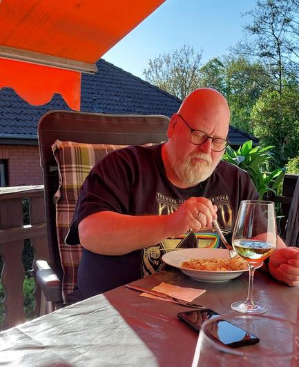 Ein Mann sitzt an einem Tisch auf einer Terrasse und genießt ein Essen. Er trägt ein schwarzes T-Shirt mit einem Aufdruck der Band Genesis und hat eine Brille auf. Vor ihm auf dem Tisch steht ein Teller mit Spaghetti, daneben ein Glas mit Weißwein. Er hält eine Gabel in der Hand und scheint das Essen zu genießen. Auf dem Tisch liegt ein Smartphone und ein Serviettenbündel. Der Hintergrund zeigt ein Gebäude mit einem dunkelgrauen Dach und grüne Bäume, die im Sonnenlicht leuchten. Eine orangefarbene Markise schützt den Tisch vor der Sonne.