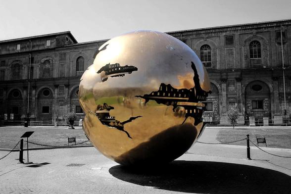 A large, polished bronze sphere with intricate cutouts stands in a paved plaza in the Vatican, reflecting the surrounding historic buildings in a partially desaturated image.