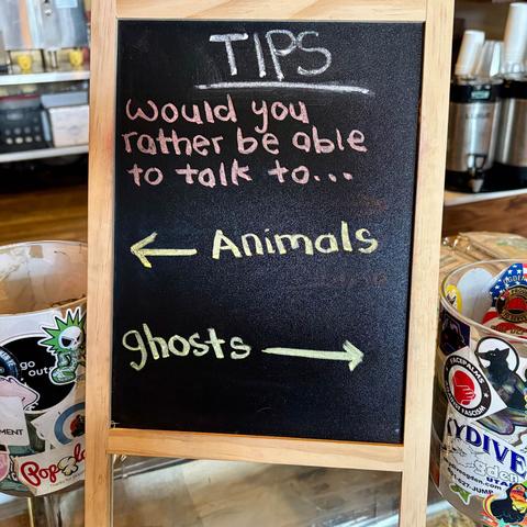 A small A-frame chalkboard sits on a counter in a coffee shop with a tip jar on either side. The chalkboard reads, "Tips. Would you rather be able to talk to…" then gives the option of "Animals" with an arrow pointing to the left just, and "Ghosts" with an arrow pointing to the right jar.