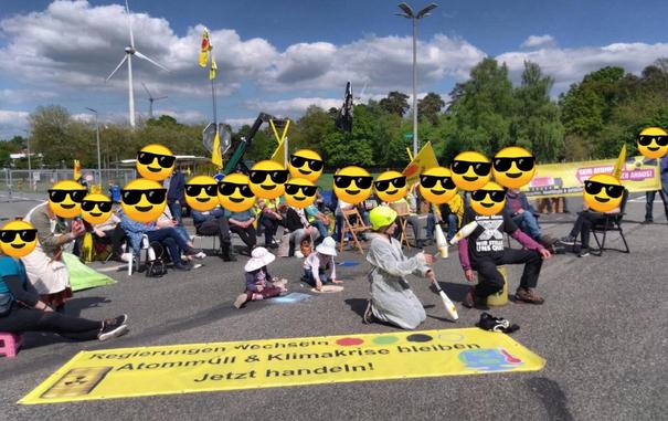 Menschen sitzen bei einer Kundgebung mit Klappstühlen, "Atommüllfässern", Fahnen und Transparenten vor dem Tor des Atommülllager Ahaus