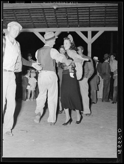 The black and white photograph captures a moment of social interaction at what appears to be an outdoor dance event. Two couples are engaged in the act of dancing; one pair is closely embraced, with the man leading by holding hands while turning his partner gently around him. The woman's posture suggests she might not be as comfortable or skilled compared to her male counterpart.

The couple behind them seems more casual and familiar with each other, possibly having a leisurely conversation rather than dancing. Their body language indicates ease in their surroundings and comfort among themselves.

In the background, there are several onlookers observing various activities around what could potentially be an open-air dance hall or community gathering spot. Some individuals appear to be children, as evidenced by one child being held aloft at a distance from the main action.

The attire of those dancing is indicative of mid-20th century fashion: men in suits and hats, women wearing dresses with stockings and high-heeled shoes. The dance floor seems informal, composed perhaps of dirt or packed earth typical for such community events during that era.

Overall, this image provides a glimpse into social customs from the early to mid 1940s at migrant camps like Shafter in California, showcasing communal gatherings where dancing was likely one popular form of entertainment. The presence of children hints at family-friendly enviro [...]