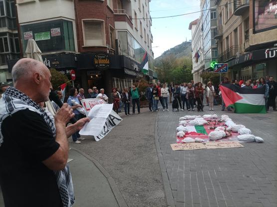 Detalle de la misma concentración donde se ve aún más gente. En primer término una persona con un pañuelo palestino al cuello lee un comunicado, micrófono en mano