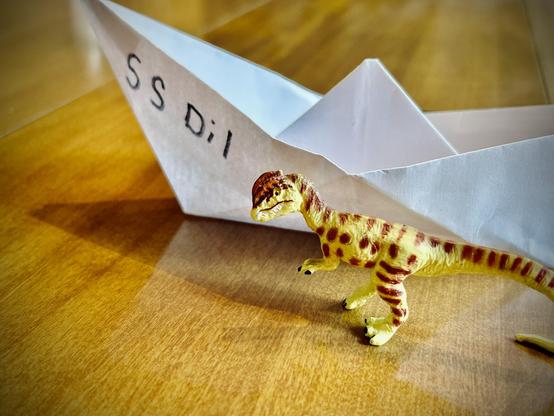 A green Dilophosaurus with brown spots stands before a white origami boat with "S.S. Dil" written on the side. Both are atop a brown dining room table.