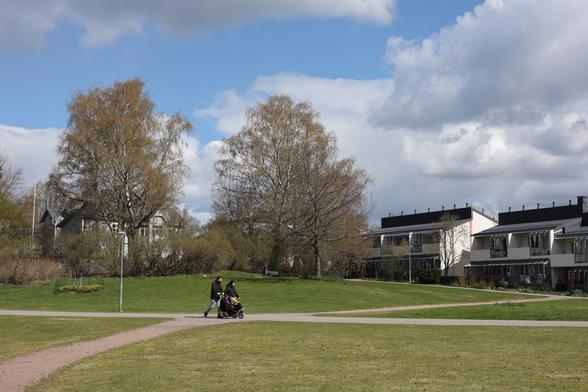 A photo of a park on a sunny spring day. There are people with a pram walking on a road.