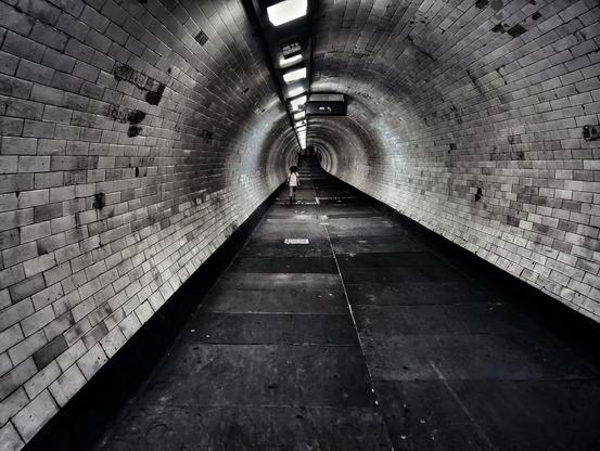 The photograph shows a long, dimly lit tunnel with a person running far in the distance. The tunnel walls are made of white, rectangular tiles, and the floor is a dark, rough surface. Overhead, there are evenly spaced lights that illuminate the tunnel's arched ceiling. The perspective of the photo emphasizes the tunnel's length, drawing the eye towards the figure at the end.