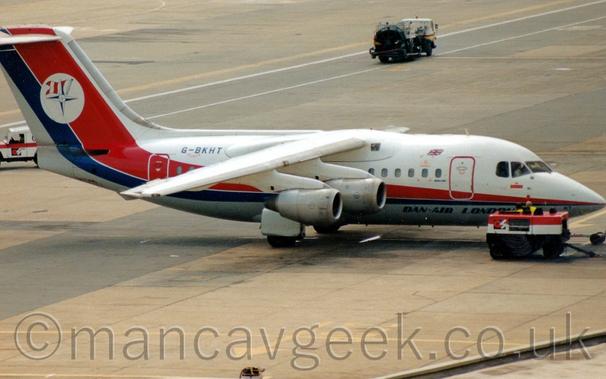 Side view of a high-winged, white, 4-engined jet airliner facing to the right but being pushed back to the left by an off-screen tug attached to the nose-wheel by a long white pole.
The plane is mostly white, with a grey belly, and a blue and red stripe running along the body, slowly getting wider as it moves rearwards, before sweeping up to fill the tail.
There are blue "Dan Air London" titles on the lower forward fuselage, and a white circle containing a white and blue compass pointer in front of a flag pole bearing a flag with vertical red and white bands.
The registration "G-BKHT" is on the upper rear fuselage in dark blue.
A red and white truck is parked in front of the nose of the plane.
Grey concrete apron fills the rest of the screen, with white lines marking out a roadway for airport vehicles, with a fuel pump in the background following the road to the top left of the screen.