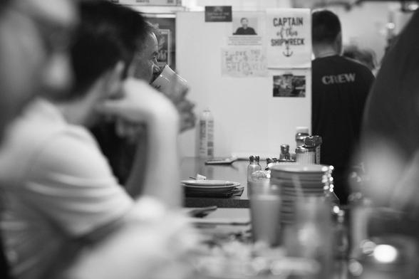 Its a long view down the countertop of a diner, selectively focused on a the man at the end pensively sipping a cup of coffee. Two other men are blurry in the foreground.