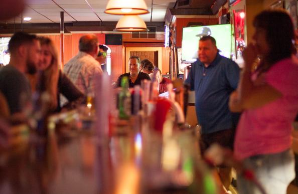 It's a view down the counter of a bar, focused on a man at the end with a bit of a smile on his face.  On the left are other customers, including a younger mixed male/female couple, both softy focused. On the right are the bartenders, also in a soft focus. This photo is lit in the warm, orange glow on the lamps and neon lights. There's a Television glowing with a baseball game, too.