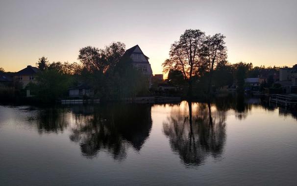 Einige Gebäude in Fürstenberg/Havel in der Abenddämmerung, einige Sonnenstrahlen spitzen zwischen fern Bäumen hindurch, vorne ein Fluss.