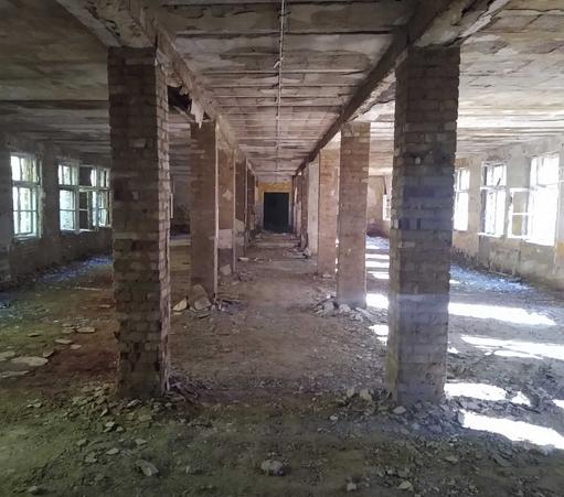 Die Innenansicht eines verfallenen Gebäudes, in der Mitte Säulen, die den Raum stützen, rechts und links offene und teils zerschlagene Fenster, am Fußboden Schutt.