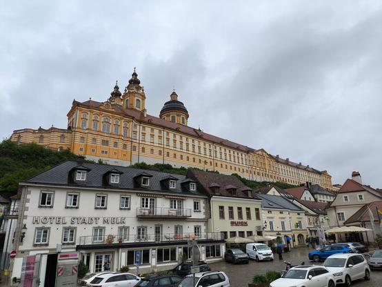 Im Vordergrund der Hauptplatz Melk mit vielen Autos und Gebäuden, ganz links mit der Aufschrift "Hotel Stadt Melk". Darüber das riesige Stiftgebäude Melk. Grauer Himmel.