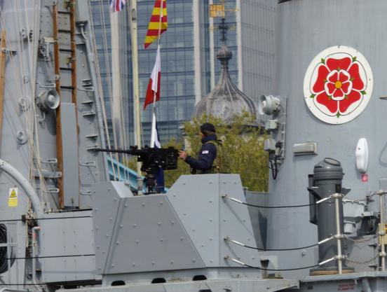 A manned heavy machine gun on the HMS Cattistock as it passed under Tower Bridge in London. The gun is black, contrasting with the matt grey of most of the ship's structure. There are a variety of flags hanging above the gun. The gunner is dressed in dark blue with a camouflaged flak jacket. The funnel behind the gunner is painted with a large red rose emblem.