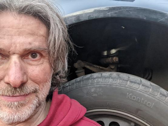 Erschöpfter älterer weißer Mann mit grauem Bart und Haaren vor einem Auto-Radkasten, aus dem 2 neue obere Querlenker hervorblitzen